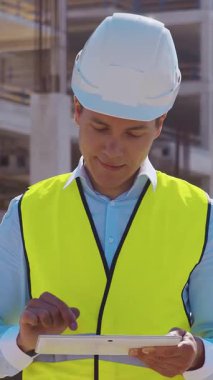 İnşaat alanının önünde duran profesyonel müteahhit. Kasklı ve yelekli ustabaşı. Ofis binası ve vinç arka planı. İş, gayrimenkul ve yatırım kavramları.