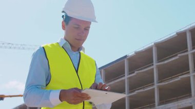 İnşaat alanının önünde duran profesyonel müteahhit. Kasklı ve yelekli ustabaşı. Ofis binası ve vinç arka planı. İş, gayrimenkul ve yatırım kavramları.