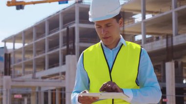 İnşaat alanının önünde duran profesyonel müteahhit. Kasklı ve yelekli ustabaşı. Ofis binası ve vinç arka planı. İş, gayrimenkul ve yatırım kavramları.