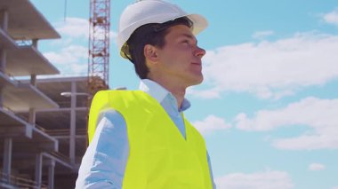 İnşaat alanının önünde duran profesyonel müteahhit. Kasklı ve yelekli ustabaşı. Ofis binası ve vinç arka planı. İş, gayrimenkul ve yatırım kavramları.