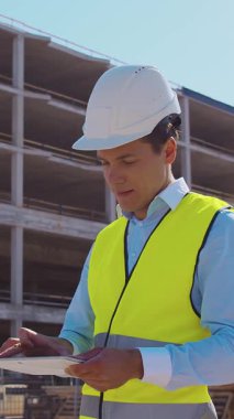 İnşaat alanının önünde duran profesyonel müteahhit. Kasklı ve yelekli ustabaşı. Ofis binası ve vinç arka planı. İş, gayrimenkul ve yatırım kavramları.