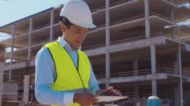 İnşaat alanının önünde duran profesyonel müteahhit. Kasklı ve yelekli ustabaşı. Ofis binası ve vinç arka planı. İş, gayrimenkul ve yatırım kavramları.