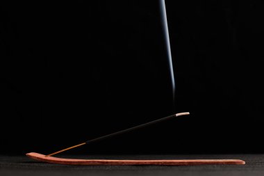 Smoke curls of incense stick in wooden holder for relaxation and meditation exercises, black background. Aromatherapy session with burning aroma stick, pleasant aroma and incense