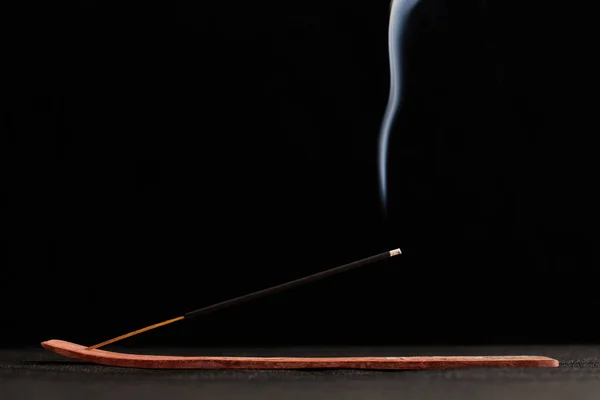 Smoke curls of incense stick in wooden holder for relaxation and meditation exercises, black background. Aromatherapy session with burning aroma stick, pleasant aroma and incense