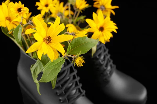 Black women platform boots with bouquet of yellow Jerusalem artichoke flowers close up, black background. Beautiful sunroot flowers in high heel platform boots, femininity shoe concept