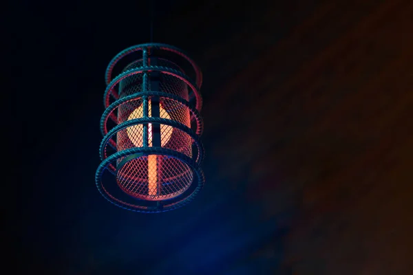 Loft pendant lamp with red blue glow in steel rebar shade, black wooden interior background, copy space. Vintage pendant lamp in modern loft cafe interior, red warm lamp bulb in steel bar shade