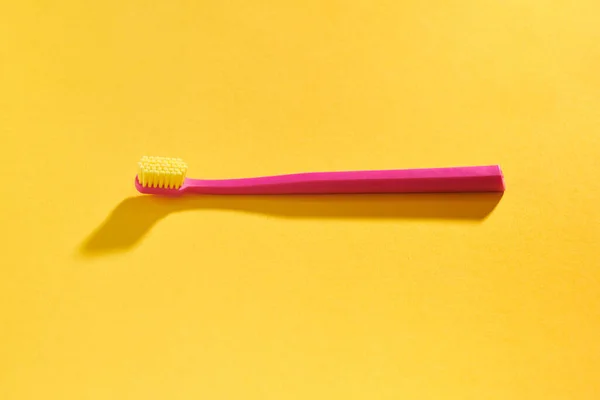 Pink toothbrush on yellow background for oral hygiene to clean teeth, gums and tongue. Manual plastic toothbrush for effective toothbrushing, hard light with shadow isolated on yellow background