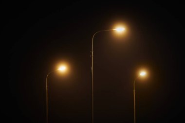 Three night lampposts shines with faint mysterious yellow light through evening fog. Streetlights shine at quiet city night, magic atmospheric light in mystical darkness
