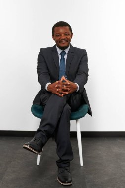 Smiling african american black man in business suit sitting on chair with crossed fingers white wall background. Full size portrait of happy black man chief executive officer waiting for interview