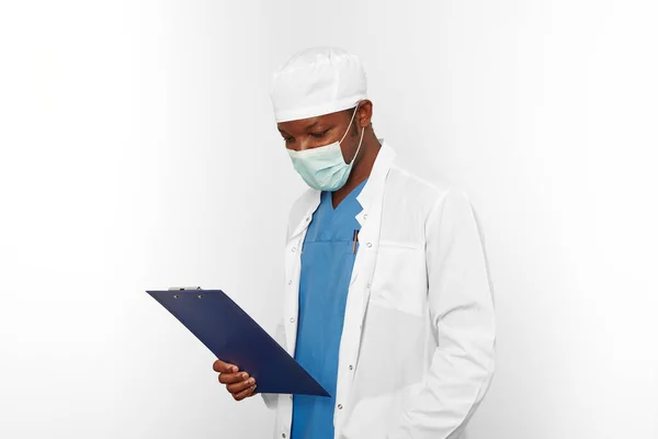 Black surgeon doctor man in white coat cap and surgeon mask looks to medical records with diagnosis on clipboard, isolated on white background. Adult black african american practicing surgeon portrait