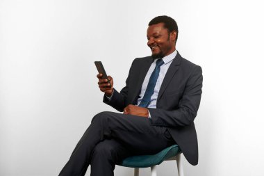 Smiling african american black man in business suit sitting on chair making video call via smartphone, white wall background. Full size portrait of happy black businessman with phone remote conference
