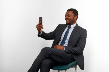 Smiling african american black man in business suit sitting on chair making video call via smartphone, white wall background. Full size portrait of happy black businessman with phone remote conference