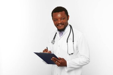 Smiling black bearded doctor man in white robe with stethoscope filling medical records on clipboard, isolated on white background. Happy adult black african american physician therapist portrait