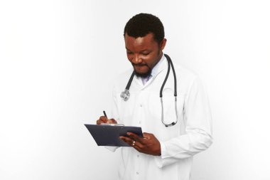Black bearded doctor man in white coat with stethoscope filling medical records on clipboard, isolated on white background. Thoughtful adult black african american physician therapist portrait