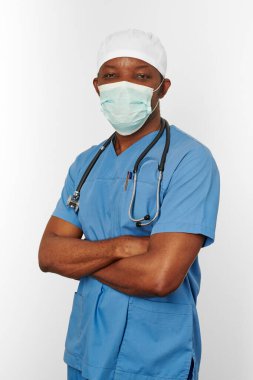 Black surgeon doctor man in blue coat white cap and surgeon mask with crossed arms isolated on white background. Focused adult black african american practicing surgeon portrait, confident look