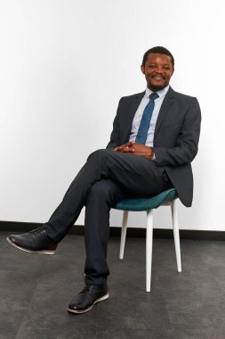 Smiling african american black man in business suit sitting on chair with crossed fingers white wall background. Full size portrait of happy black man chief executive officer waiting for interview