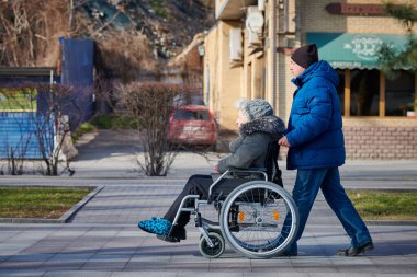 Rostov on Don, Russia - 02.18.2022 - Adult man rolls his disabled mother on wheelchair, careful relationships with disabled person. Adult woman health recovery and rehabilitation after stroke
