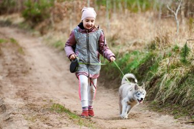 Girl walking on leash Siberian Husky puppy on country road at forest background, happy friendship of dog and cute little kid. Young girl running Husky dog breed puppy on autumn outdoor road