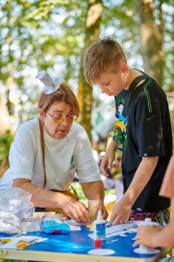 Svetlogorsk, Russia - 13.08.2022 - Funny adult woman in small paper hat teaches young guy make paper craft paper and scrapbooking at outdoor art festival, handmade female teacher for children