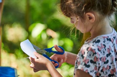 Svetlogorsk, Russia - 13.08.2022 - Little girl cuts piece of paper with scissors at outdoor art paper craft lesson, teaching children to create paper crafts. Little lady passionate about her hobby