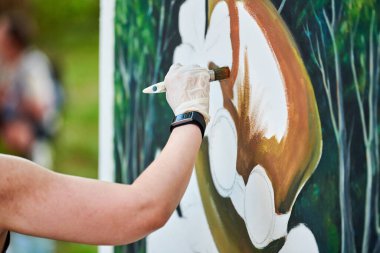 Ressam kız elinde boya fırçasıyla açık hava resim festivalinde beyaz tuvale yeşil bir manzara çiziyor, resim çiziyor. Kadın sanatçı atmosferik gerçeküstü resim çiziyor.