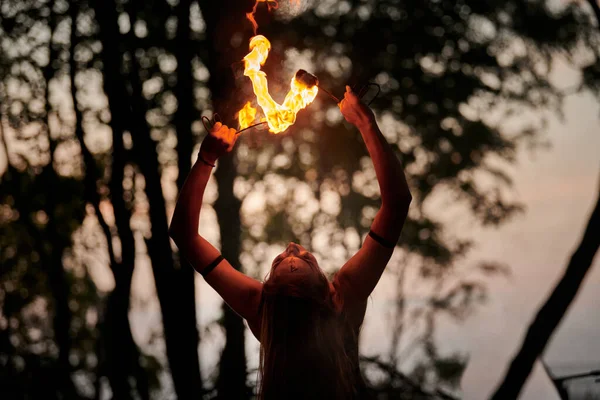 Girl fire dancing performance at outdoor art festival, smooth movements of female fire show artist with torch. Woman dancing with fire flame art performance, twilight forest background