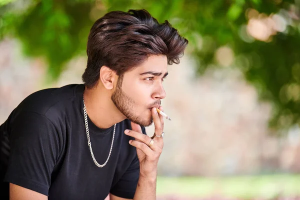 Young indian man smoker portrait in black t shirt and silver neck chain in public park, hindu male smoking close up portrait. Handsome indian man portrait with thick hair in city green park