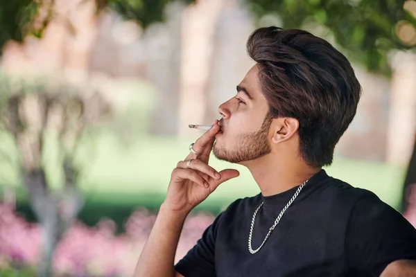 Young indian man smoker portrait in black t shirt and silver neck chain sitting on bench in public park, hindu male smoking close up portrait. Handsome indian man portrait with thick hair in city park