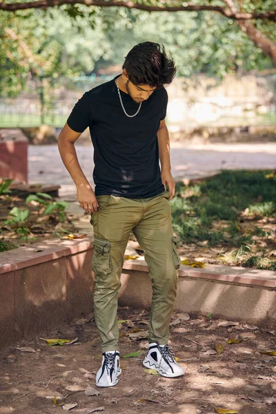 Attractive young indian man full height portrait in black t shirt and silver neck chain in green park, hindu male portrait. Handsome indian man outdoor portrait with thick hair in public park