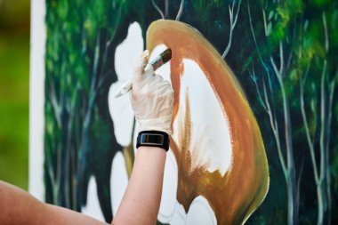 Ressam kız elinde boya fırçasıyla açık hava resim festivalinde beyaz tuvale yeşil bir manzara çiziyor, resim çiziyor. Kadın sanatçı atmosferik gerçeküstü resim çiziyor.