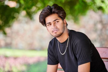 Çekici Hintli sigara içen adam siyah tişörtle sigara dumanı portresi ve halka açık parkta gümüş boyun zinciri veriyor. Hindu erkek, yakın portre sigara içiyor. Kalın saçlı, yakışıklı Hintli adam portresi.