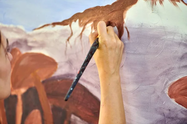 Girl artist hand holds paint brush and draws abstract surreal landscape on white canvas at outdoor art painting festival, paintings art picture process. Woman artist paints atmospheric surreal picture