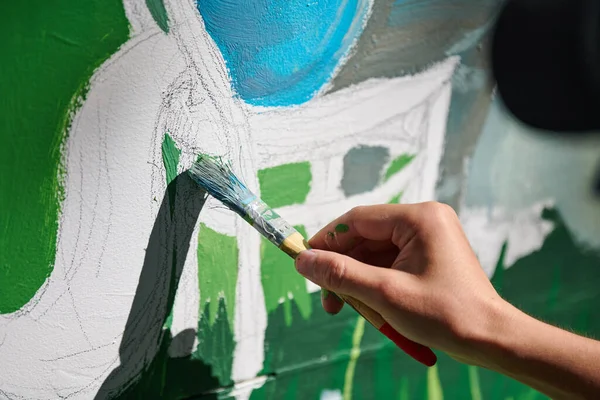 Girl artist hand holds paint brush and draws green nature landscape on white canvas at outdoor art painting festival, paintings art picture process. Woman artist paints atmospheric surreal picture