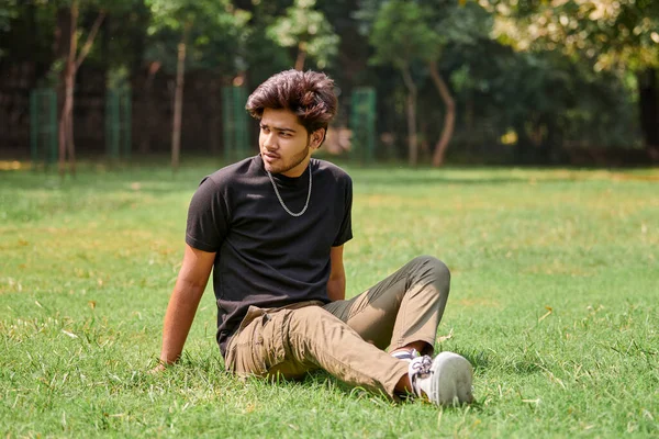 Attractive young indian man portrait in black t shirt and silver neck chain sitting on green lawn in public park, hindu male portrait. Handsome indian man portrait with thick hair in city park