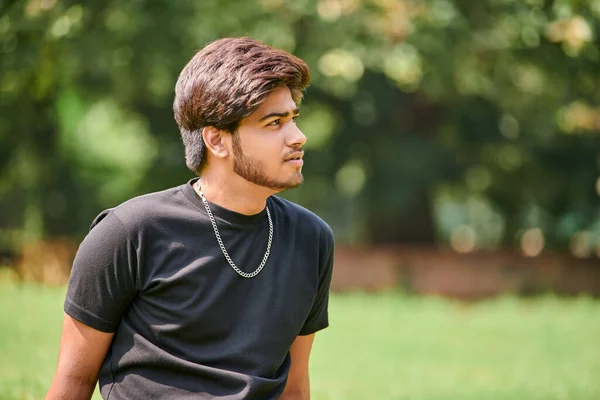 Attractive young indian man portrait in black t shirt and silver neck chain sitting on green lawn in public park, close up hindu male portrait. Handsome indian man portrait with thick hair in park