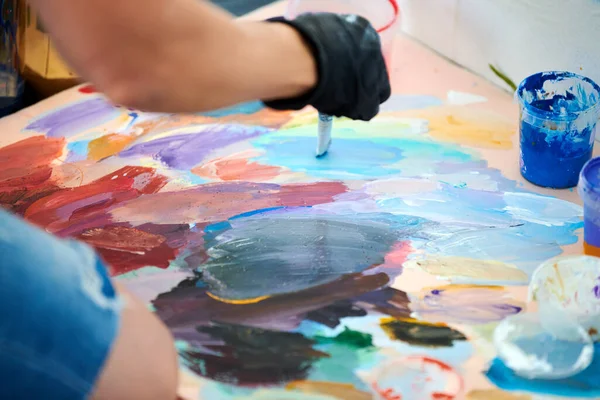 Artist hand in black gloves holds paint brush and choose color from colorful palette at outdoor art painting festival, paintings art picture process. Artist paints atmospheric surreal picture