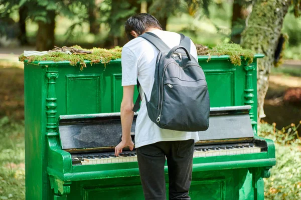 Stock image Young guy teenager plays classic music on piano, interactive outdoor art object in public park, rear view. Young man in casual clothes with backpack plays piano in green forest, unity with nature