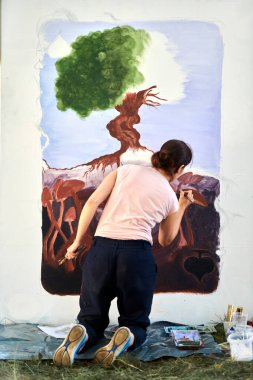 Kız ressam, boya fırçasıyla doğa üstü bir manzara çizer açık hava resim festivalinde beyaz tuval üzerine resim, resim süreci, dikiz manzarası. Kadın sanatçı atmosferik gerçeküstü resim çiziyor.
