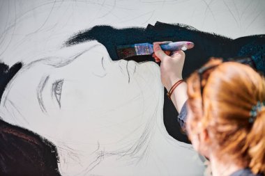 Kadın sanatçı, açık hava resim festivalinde beyaz tuvale boya fırçası sürrealist kız portresi çiziyor, resim çiziyor. Siyah şapkalı kadın atmosferik gerçeküstü resim çiziyor.