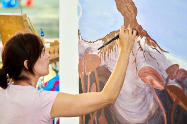 Kız ressam, boya fırçasıyla doğa üstü bir manzara çizer açık hava resim festivalinde beyaz tuval üzerine resim, resim süreci, dikiz manzarası. Kadın sanatçı atmosferik gerçeküstü resim çiziyor.