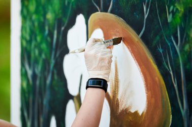 Ressam kız elinde boya fırçasıyla açık hava resim festivalinde beyaz tuvale yeşil bir manzara çiziyor, resim çiziyor. Kadın sanatçı atmosferik gerçeküstü resim çiziyor.