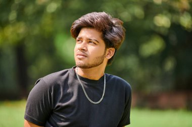 Siyah tişörtlü çekici genç Hintli adam portresi ve açık yeşil park arka planında gümüş boyun zinciri, Hindu erkek portresini kapat. Halk parkında gür saçlı yakışıklı Hintli adam portresi