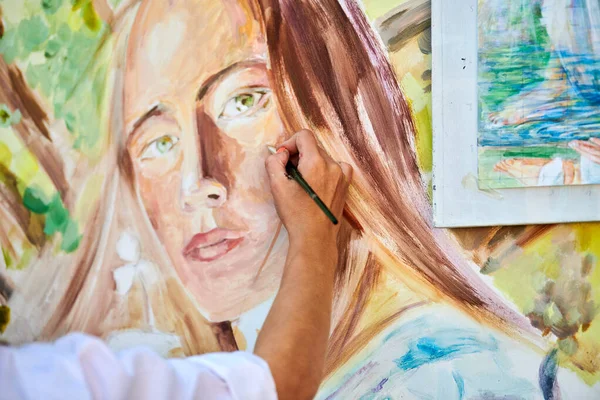 Girl artist hand holds paintbrush and draws surreal woman portrait on white canvas at outdoor art painting festival, paintings art picture process. Woman artist paints atmospheric surreal picture