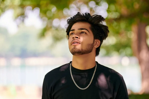 Attractive young indian man portrait in black t shirt and silver neck chain on outdoor green park background, close up hindu male portrait. Handsome indian man portrait with thick hair in public park