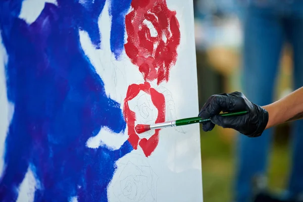 Artist hand in black gloves holds paint brush and draws abstract surreal image on white canvas at outdoor art painting festival, paintings art picture process. Artist paints atmospheric picture