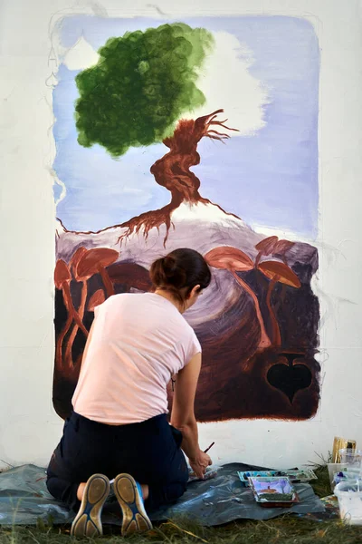 Girl artist draws with paint brush surreal nature landscape on white canvas at outdoor art painting festival, paintings art picture process, rear view. Woman artist paints atmospheric surreal picture