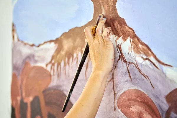 Girl artist hand holds paint brush and draws abstract surreal landscape on white canvas at outdoor art painting festival, paintings art picture process. Woman artist paints atmospheric surreal picture