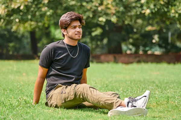公共公園で緑の芝生の上に座って黒のTシャツと銀の首のチェーンで魅力的な若いインドの男性の肖像画 Hindu男性の肖像画 都市公園で厚い髪を持つハンサムなインド人の男性の肖像画 — ストック写真