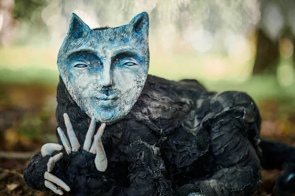 Black cat in human face mask art object at outdoor art exhibition in public park, weird black creature in mask lying on autumn foliage. Creepy strange cat art object, duplicity and fallacy concept