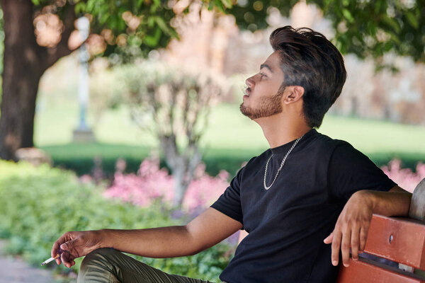 Attractive indian man smoker exhales cigarette smoke portrait in black t shirt and silver neck chain sitting on bench in public park, hindu male smoking close up portrait. Handsome indian man portrait